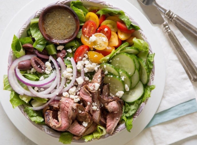 Flank steak salad with onion, olives, cucumbers, peppers, and dressing plated in a salad bowl.