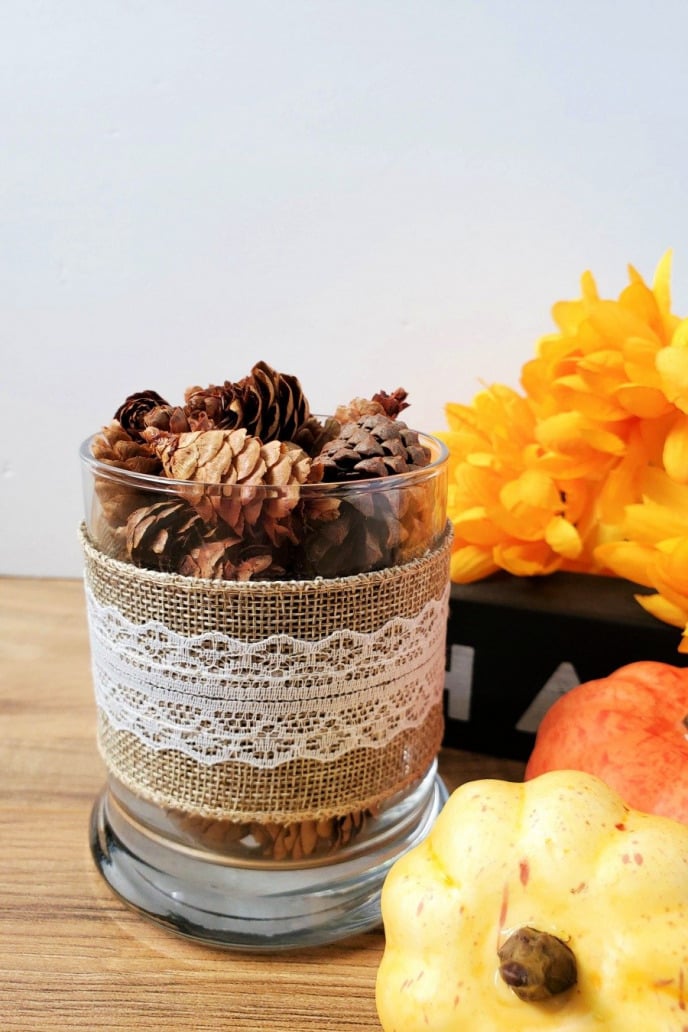 Pine cone craft decor, pine cones in a glass jar wrapped in burlap sitting next to pumpkins and fall flowers