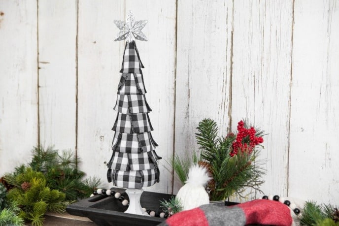 Buffalo plaid Christmas decoration, black and white buffalo plaid ribbon tree on a white tree stand, flanked by decorations like holly and twigs.
