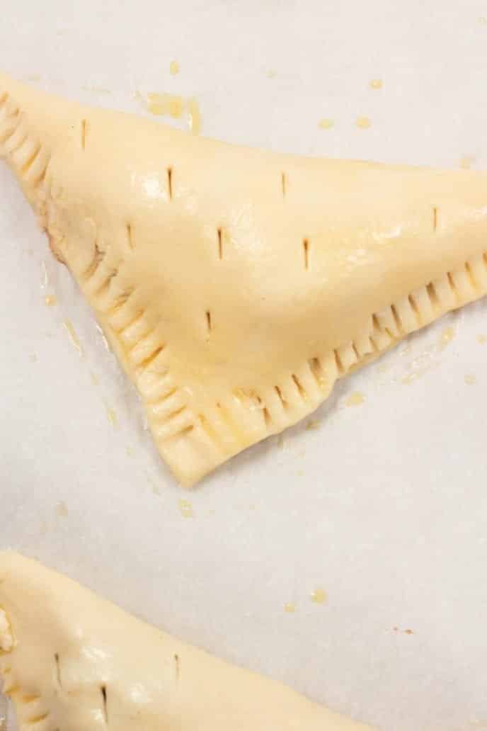 Apple turnover dough filled and folded into a triangle with the edges crimped, egg wash, and pierced for steam release while baking.
